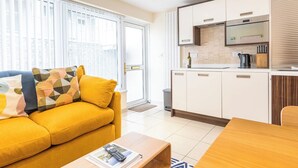 Un appartement moderne avec un canapé jaune, une table à manger en bois, une cuisine dotée de meubles blancs et une grande fenêtre avec des stores.