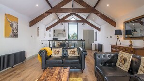 A spacious living room with a high ceiling, exposed wooden beams, a black leather sofa set, and a view into the kitchen area.