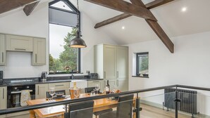 A spacious kitchen with high ceilings and exposed beams, a dining area with a wooden table and chairs, and a view of greenery outside.