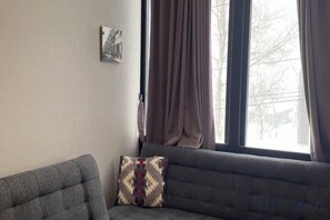 A room with a grey sofa, a patterned pillow, a window with curtains, and a wall-mounted picture. - Q Fox Black Diamond Hotel (Kutchan)