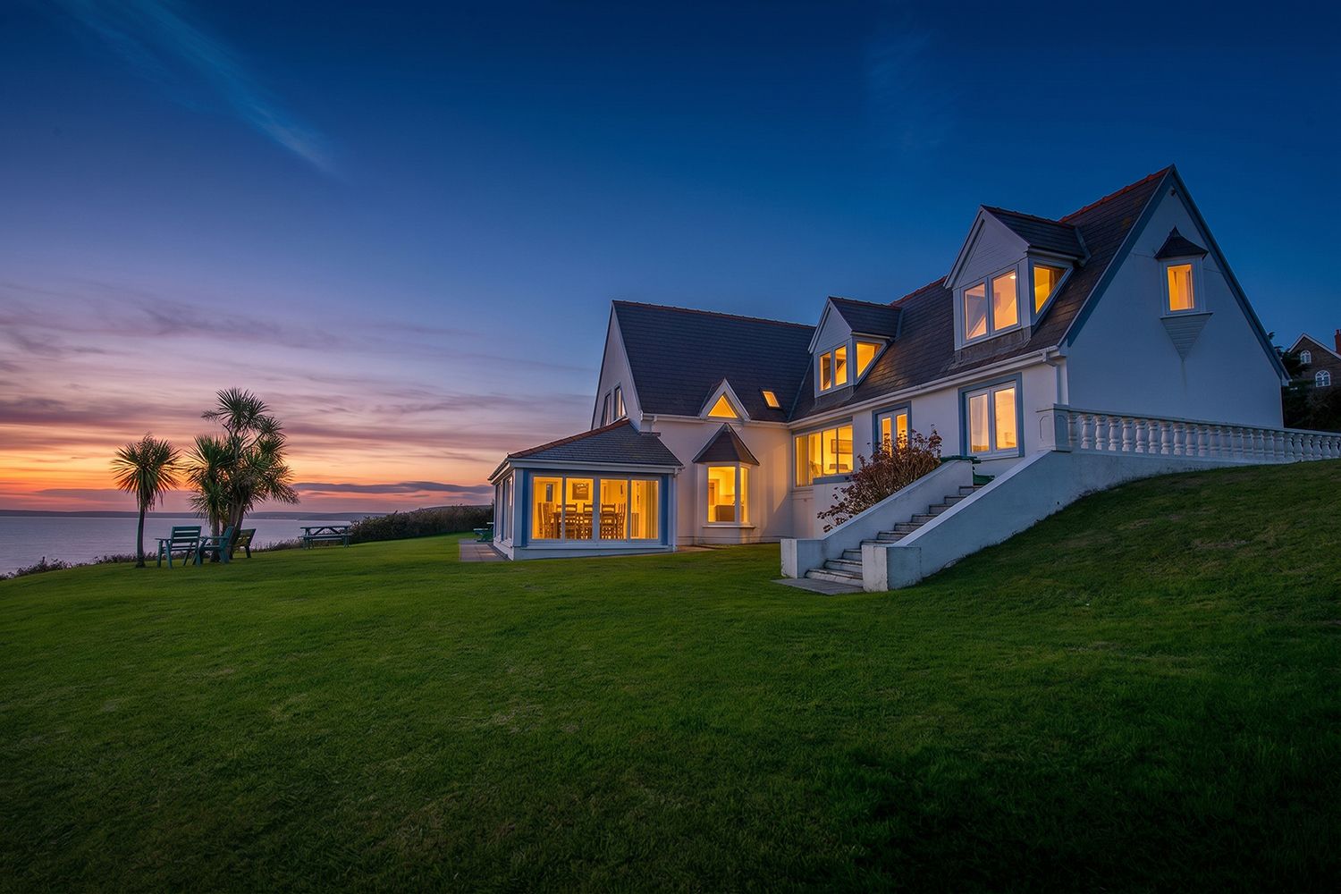A large house with a well-maintained lawn, a staircase leading to the entrance, and a view of the sea at sunset.