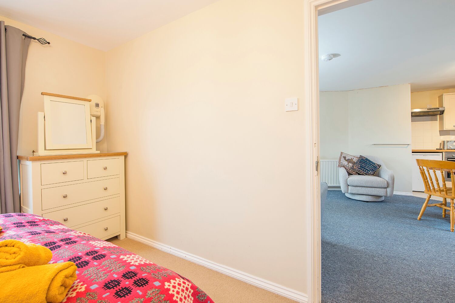 A bedroom with a bed, a dresser, a mirror, and a door leading to a living area with a chair and a table.
