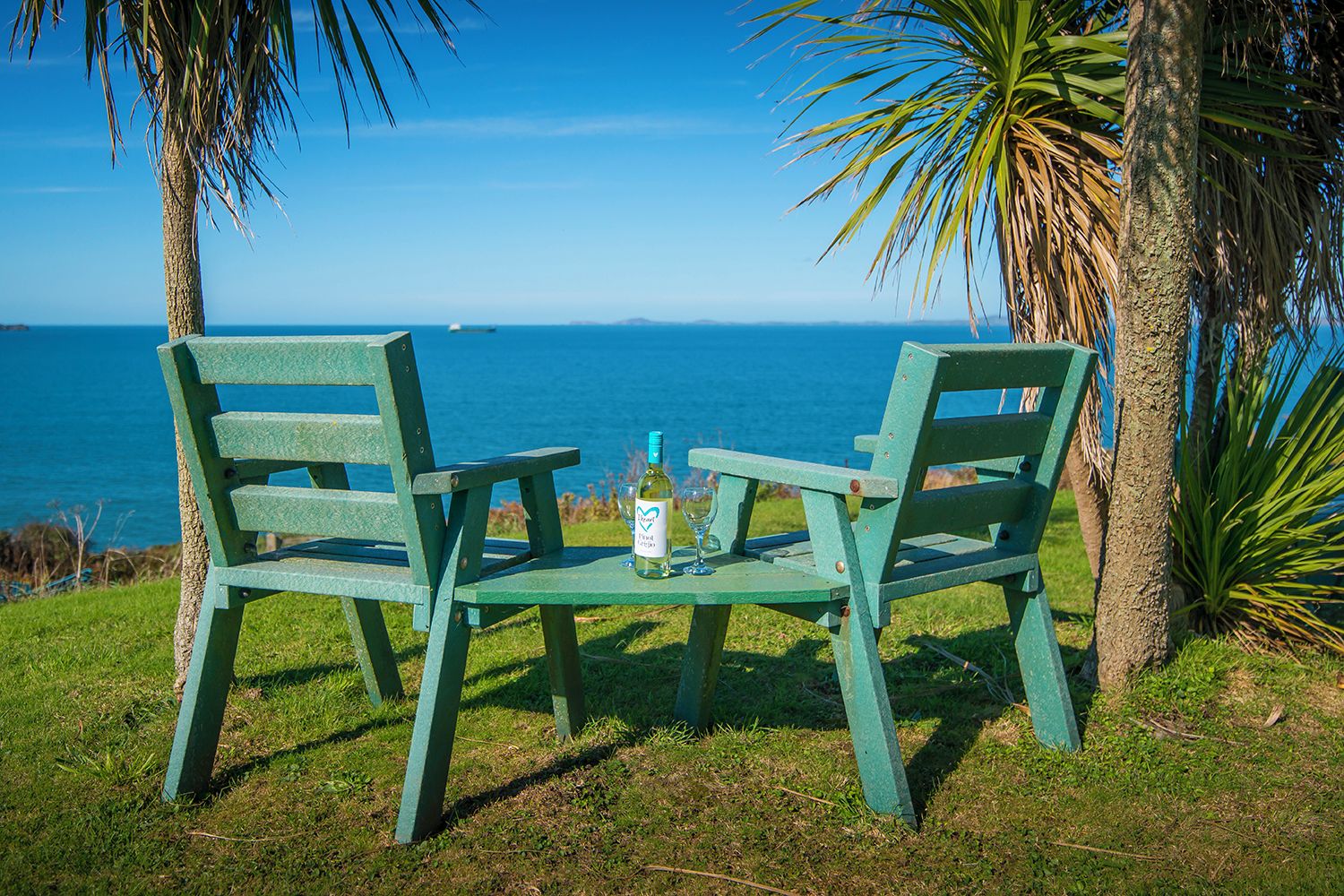 Two green chairs facing a body of water with a bottle and glass on the table.