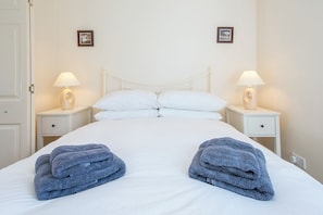 A neatly made bed with white linens and two folded towels on each side. There are two bedside tables with lamps and a small drawer. The walls are plain, and there are two framed pictures on the wall.