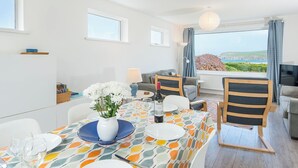 Uma sala de jantar com uma mesa posta para quatro pessoas, um sofá e uma televisão, com vista para o mar.