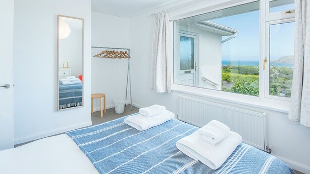 Quarto com uma janela grande, cama com roupa de cama listrada em azul, mesa de cabeceira, espelho e vista para o verde e o mar.