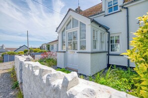 Une maison blanche dotée d’une grande baie vitrée, d’un mur en pierre et d’un jardin où poussent différentes plantes et fleurs.