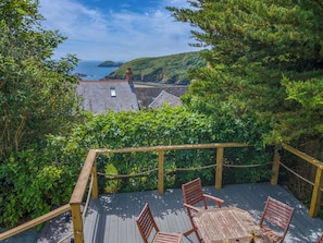 A wooden deck with chairs and a table, overlooking a coastal landscape with hills and a view of the sea.