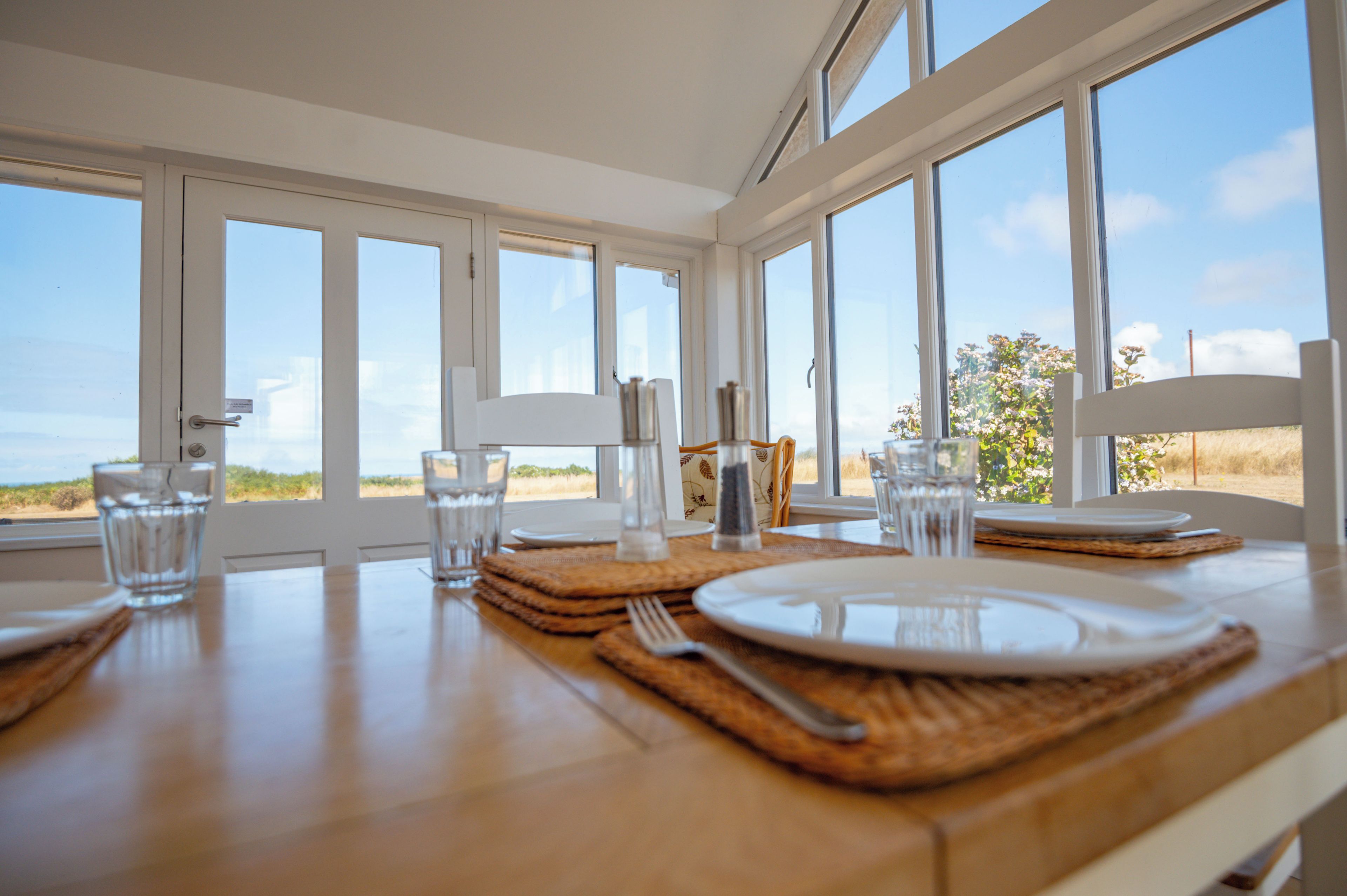 Una mesa de comedor dispuesta con platos, vasos y cubiertos, con vista al exterior a través de amplios ventanales.