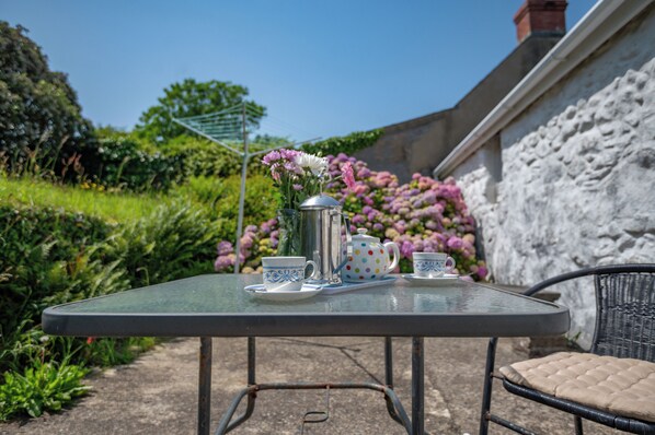 A table with a teapot, cups, and saucer set, positioned outdoors with a garden and a stone wall in the background. - Bwthyn Bach, Holiday Cottage Trefin, Sleeps 2, 1 bedrooms, bathrooms (Haverfordwest)