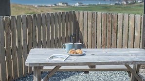 Et træbord med et picnic-setup: to kopper og et fad med kager, foran et træhegn med et naturskønt udsyn i baggrunden.