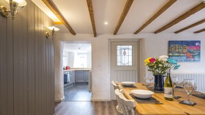 A dining area with a wooden table, chairs, and a vase of flowers. There is a kitchen in the background with a window and a painting on the wall. - Carew Cottage, Holiday Cottage Carew, Sleeps 4, 2 bedrooms,  bathrooms (Tenby)