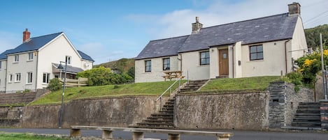 A coastal building with a stone staircase leading to a wooden door, a picnic table in the foreground, and a clear blue sky.