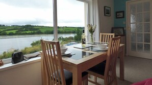 Uma área de refeições com vista para um rio, uma mesa posta com uma tigela, garrafa de vinho e copos, e duas cadeiras de madeira.