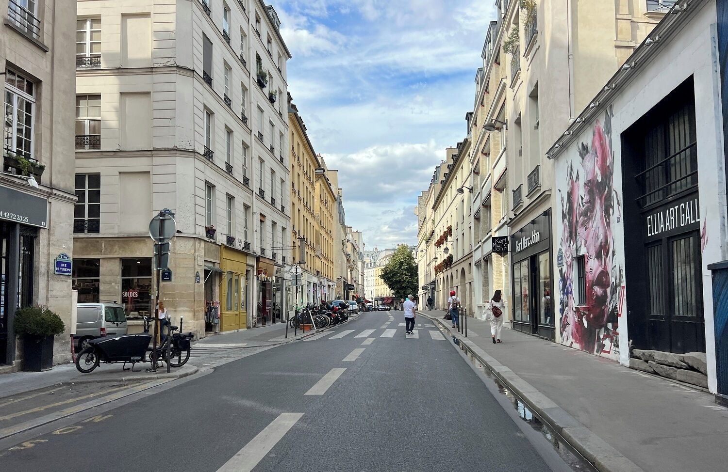 A street view with buildings on both sides, a clear sky, and a few pedestrians.