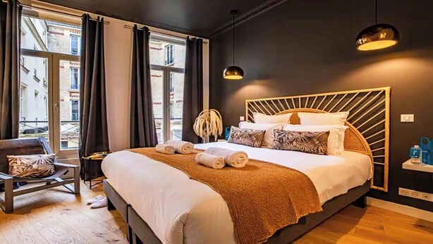 A modern hotel room with a large bed, a chair, a small table, and a view of the city through the window.