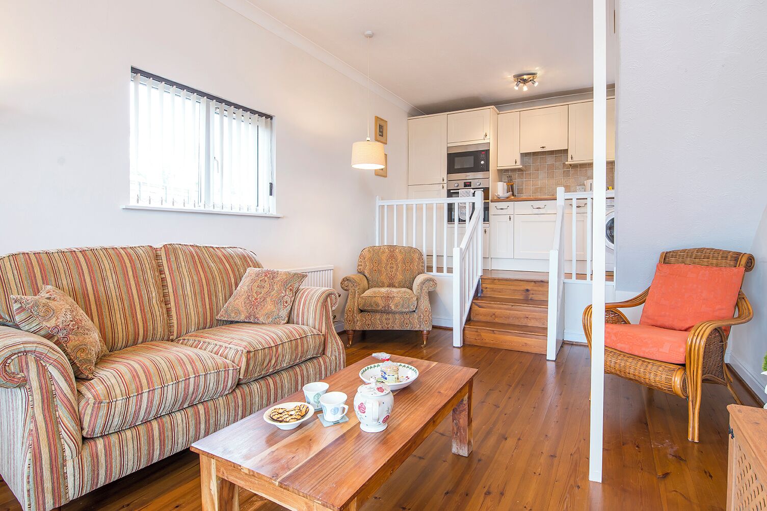 Una sala de estar con un sofá a rayas, un sillón estampado, una mesa de centro de madera con tazas de té y una silla de mimbre con un cojín naranja. Se ve una cocina al fondo, con armarios blancos y una escalera de madera.