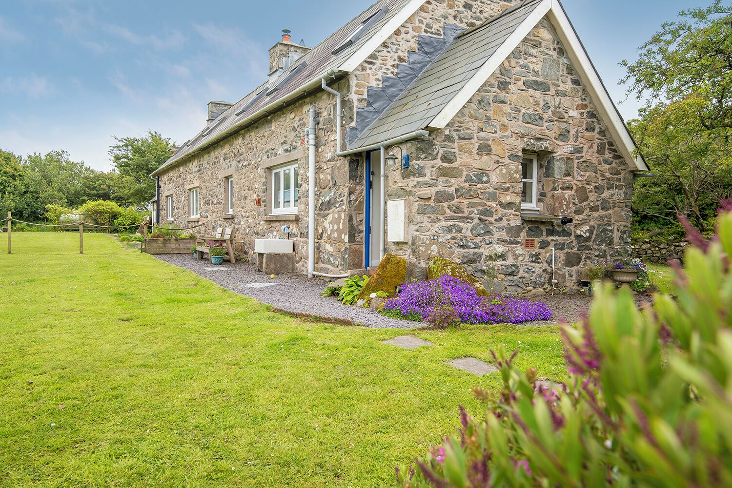 A stone house with a sloped roof, a front door, and a small garden with purple flowers.