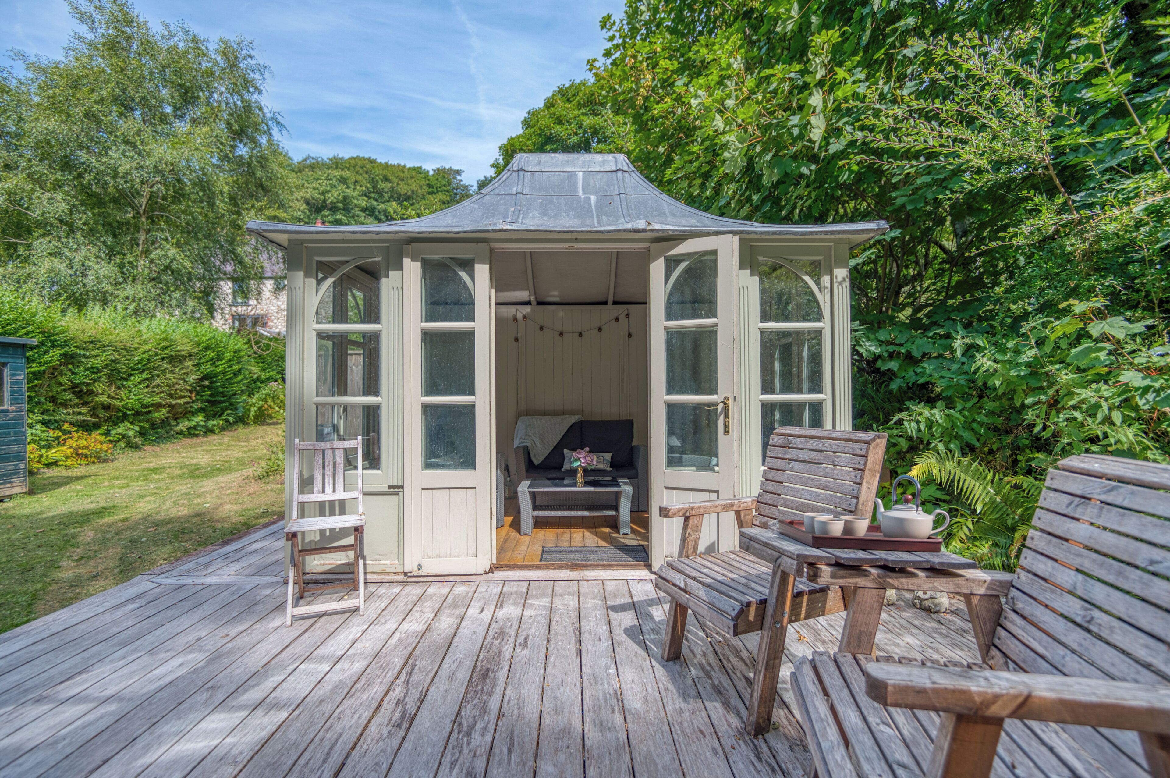 A wooden deck with a gazebo-style room featuring a sofa, chairs, and a table with tea set.