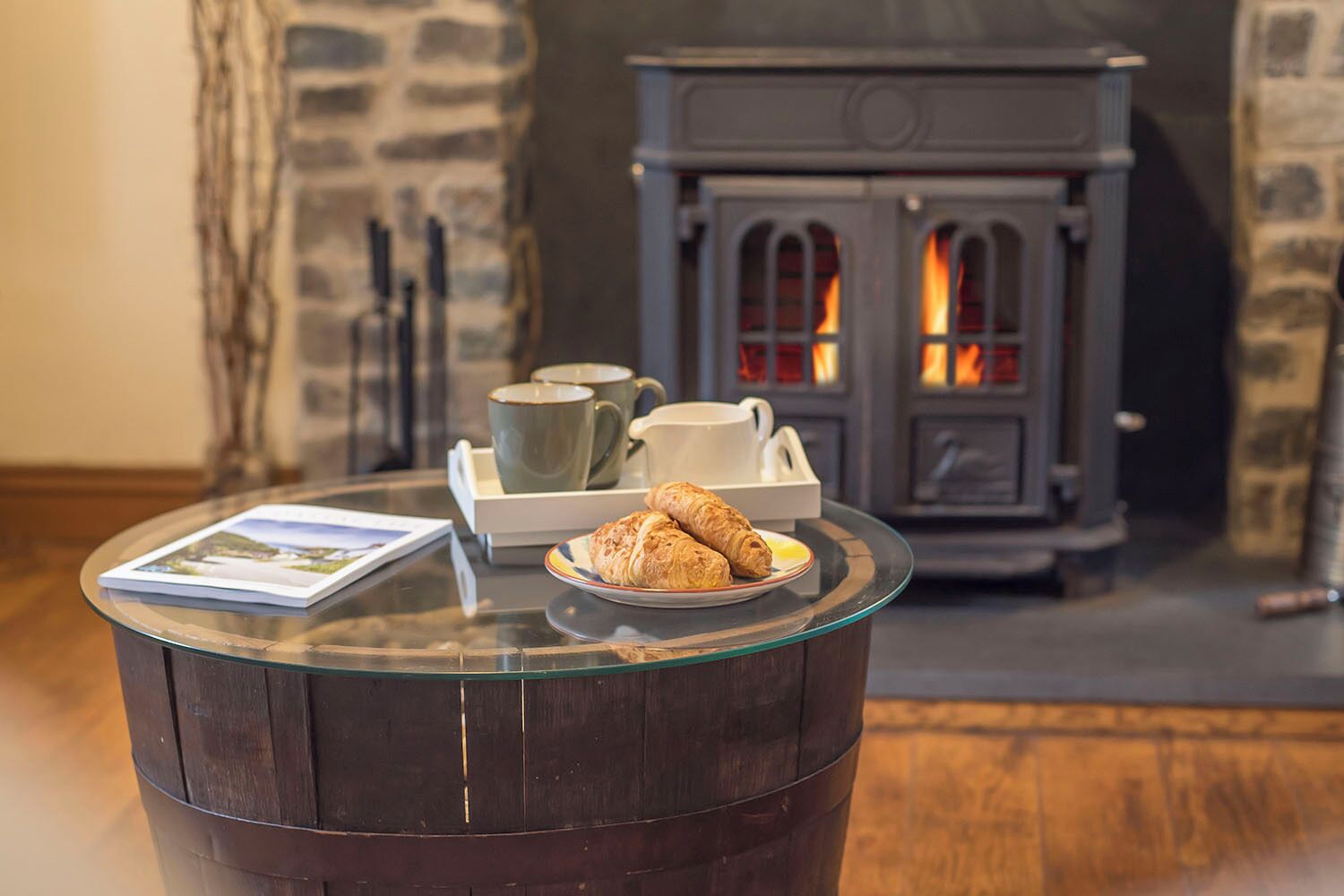 A cozy room with a glass table displaying a croissant, a book, and a tray with cups and saucers. A stone fireplace with a visible fire adds warmth to the scene.