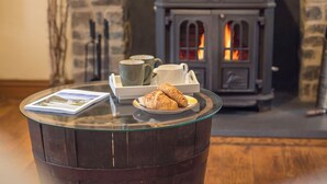 A cozy room with a glass table displaying a croissant, a book, and a tray with cups and saucers. A stone fireplace with a visible fire adds warmth to the scene. - Fairholme, Holiday Cottage Solva, Sleeps 4, 2 bedrooms,  bathrooms (Haverfordwest)