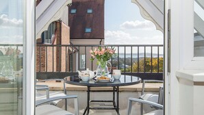 A balcony with a table set for tea, a vase with flowers, and a view of buildings and hills.