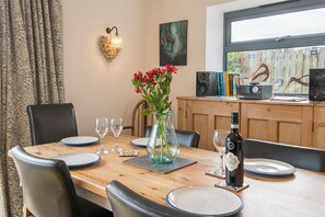A dining table set with plates, glasses, a vase with flowers, a bottle of wine, and a wall-mounted light fixture. - Glan y Mor, Holiday Home Freshwater East, Sleeps 6, 3 bedrooms, bathrooms (PEMBROKE)