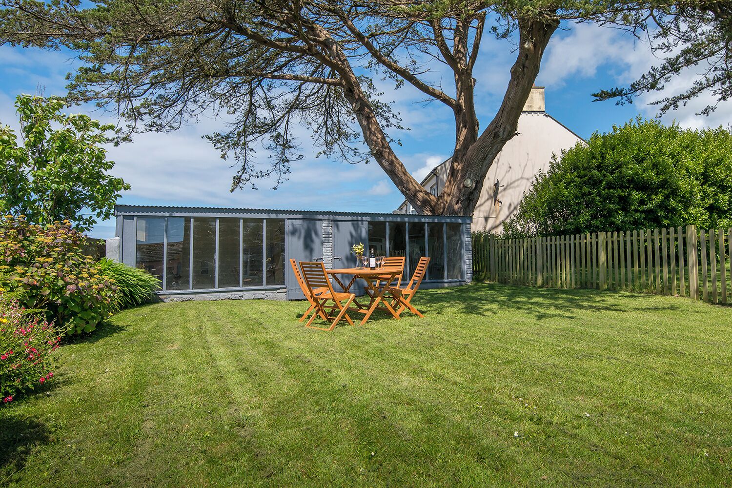 A backyard with a wooden table and chairs, a large tree, and a glass-enclosed structure.