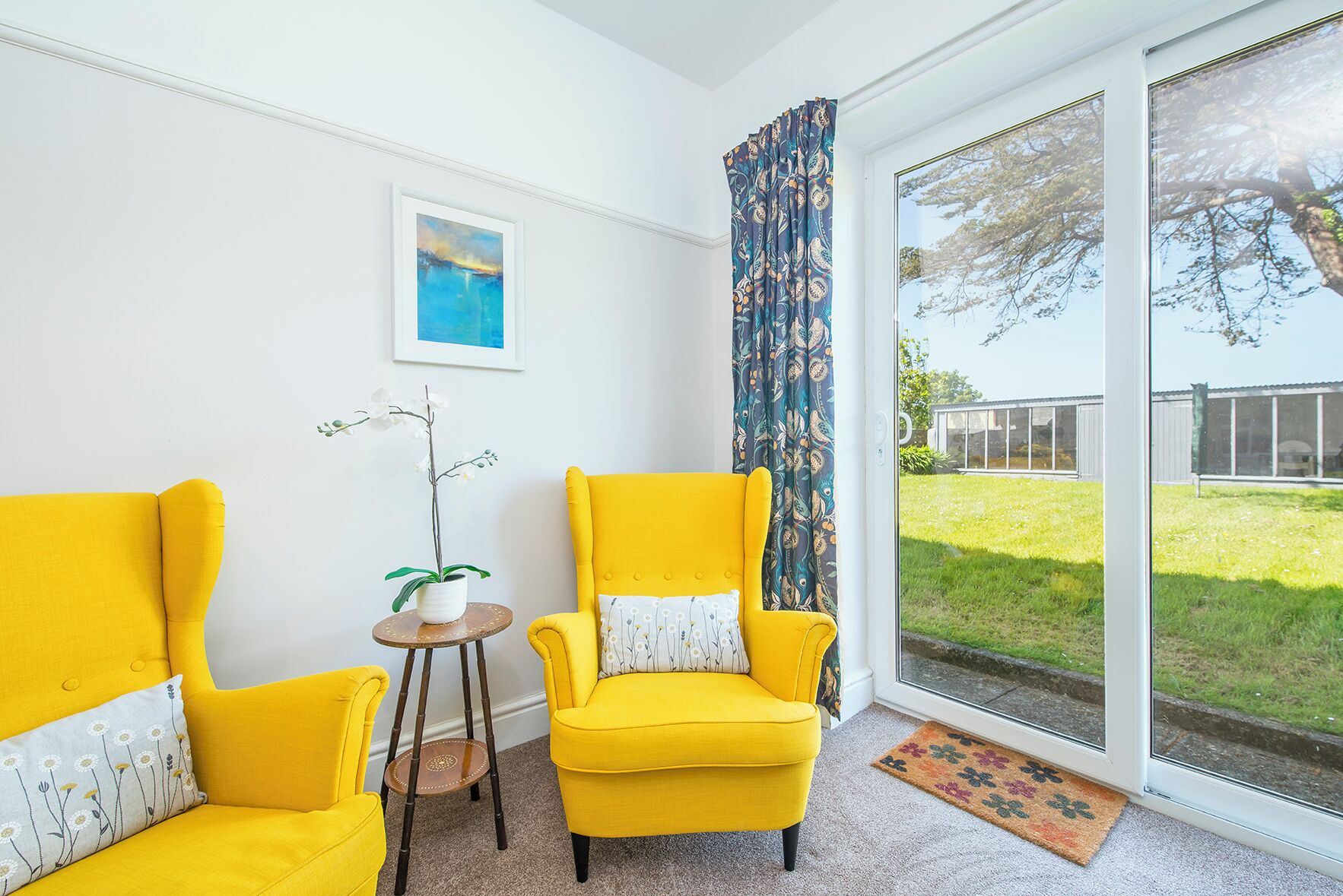 A room with two yellow armchairs, a small table with a plant, a framed picture on the wall, and a large sliding glass door leading to a grassy outdoor area.