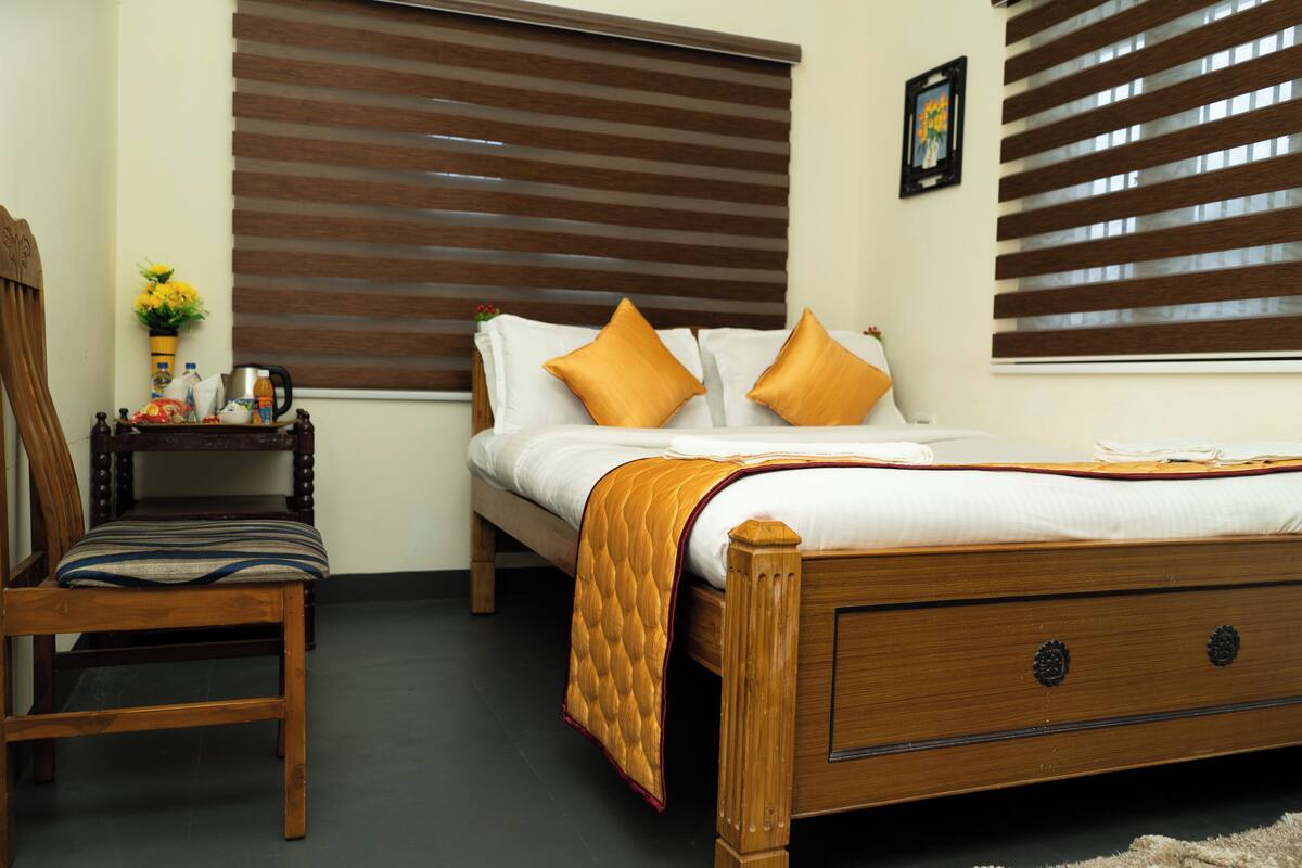 A bedroom with a wooden bed, two pillows, a bedside table with a lamp, a chair, and a window with blinds.