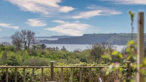 A coastal view with a wooden deck and a table, overlooking a body of water and cliffs. - Green Plains, holiday home Wisemans Bridge, Sleeps 6, 3 bedrooms,  bathrooms (Narberth)