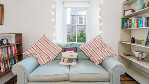 A cozy living room with a light blue sofa, striped pillows, a bookshelf, and a window with a view of a building. - Greystones House, Holiday Home St Davids, Sleeps 6, 3 bedrooms,  bathrooms (Haverfordwest)