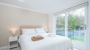 Ein modernes Schlafzimmer mit einem großen Bett, weißer Bettwäsche, einem Nachttisch mit Lampe und Blick auf einen Garten durch eine Schiebetür.