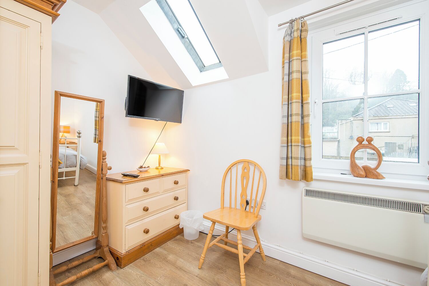 A bedroom with a skylight, a television, a wooden dresser, a chair, and a window with curtains.
