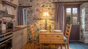 A rustic kitchen with a stone wall, wooden cabinets, a stove, and a dining table with chairs. - Heather Cottage, Holiday Home Near Nolton Haven, Sleeps 2, 1 bedrooms,  bathrooms (Portfield Gate)