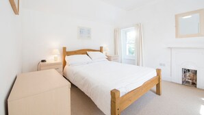 A bedroom with a wooden bed, bedside tables with lamps, a small cabinet, a fireplace, and a window with curtains. - Inglenook, Holiday Cottage Manorbier, Sleeps 4, 2 bedrooms,  bathrooms (Manorbier)