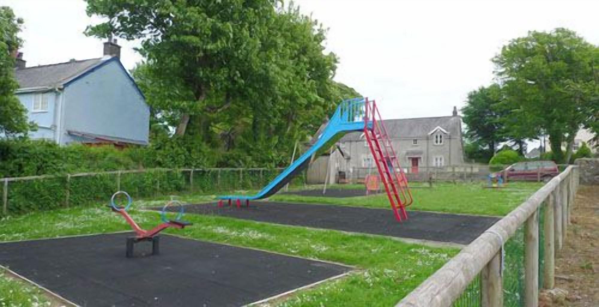 A playground with a blue slide and red climbing structure, surrounded by a fence and greenery.