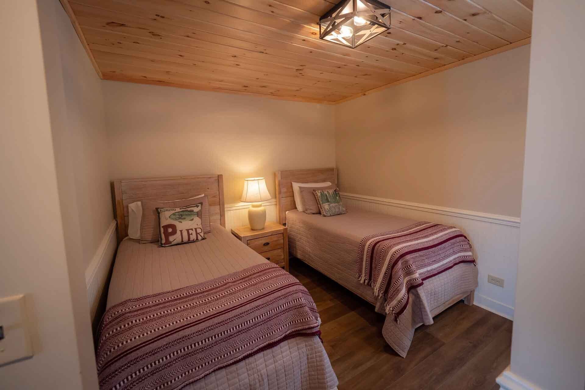 A small, cozy bedroom with two single beds, wooden ceiling, and a nightstand with a lamp.