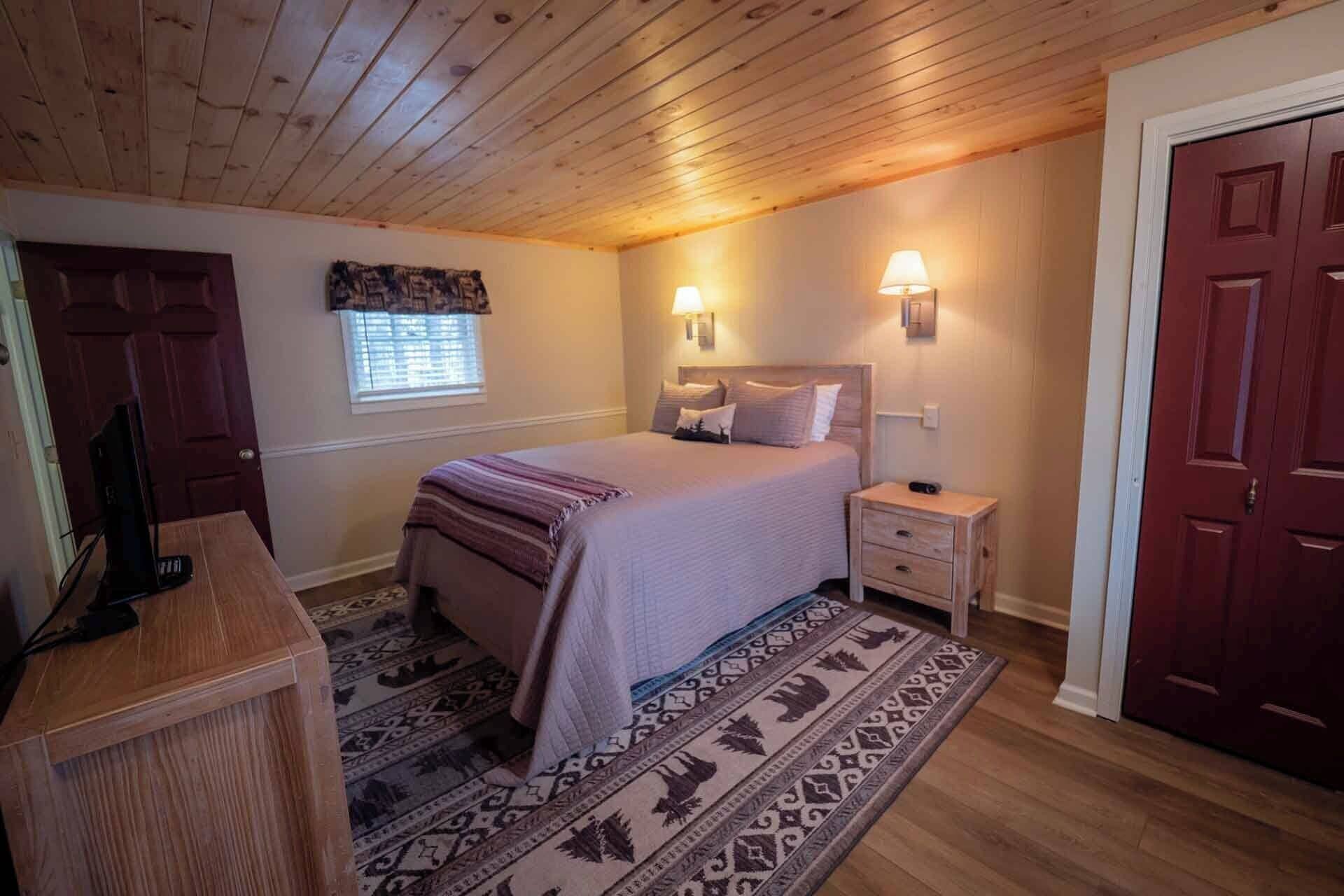 Un dormitorio con cama, lámparas de noche, un escritorio de madera, una alfombra estampada y una puerta.