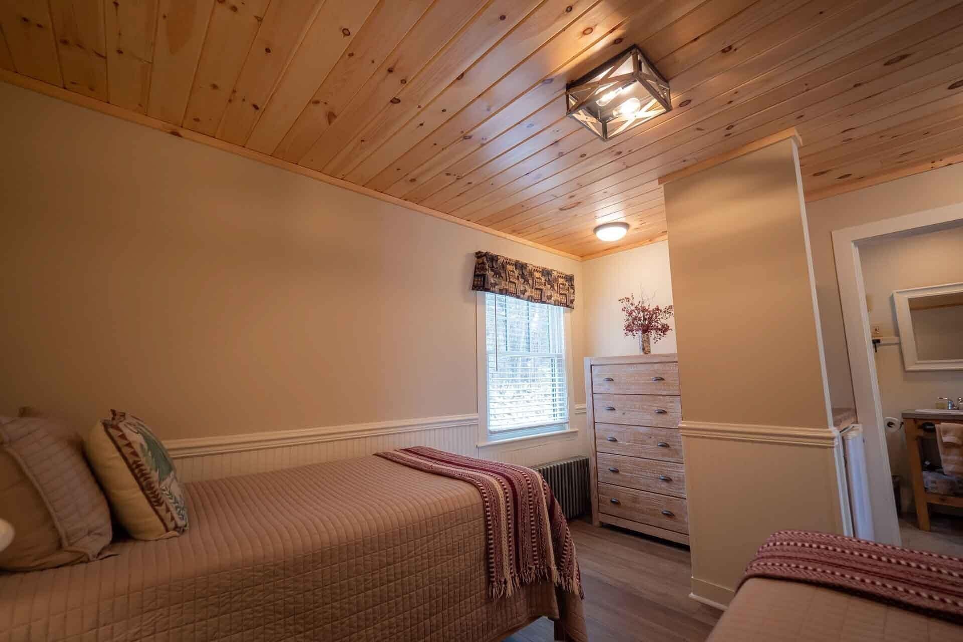 A bedroom with a wooden ceiling, a window with blinds, a dresser, and a bed with a quilted blanket.