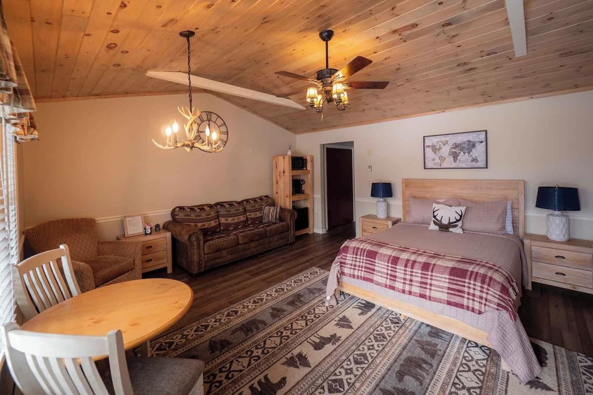 Een gezellig slaapkamer met een houten plafond, een bed met een geruit plaid, een houten hoofdbord, een nachtkastje met een lamp, een houten bank, een kamerbreed tapijt met een patroon, en een kroonluchter.