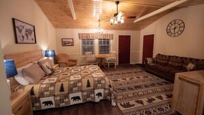 Une pièce chaleureuse avec un plafond en bois, un lit recouvert d’une courtepointe à motifs, un canapé, une table à manger avec des chaises et une horloge au mur.