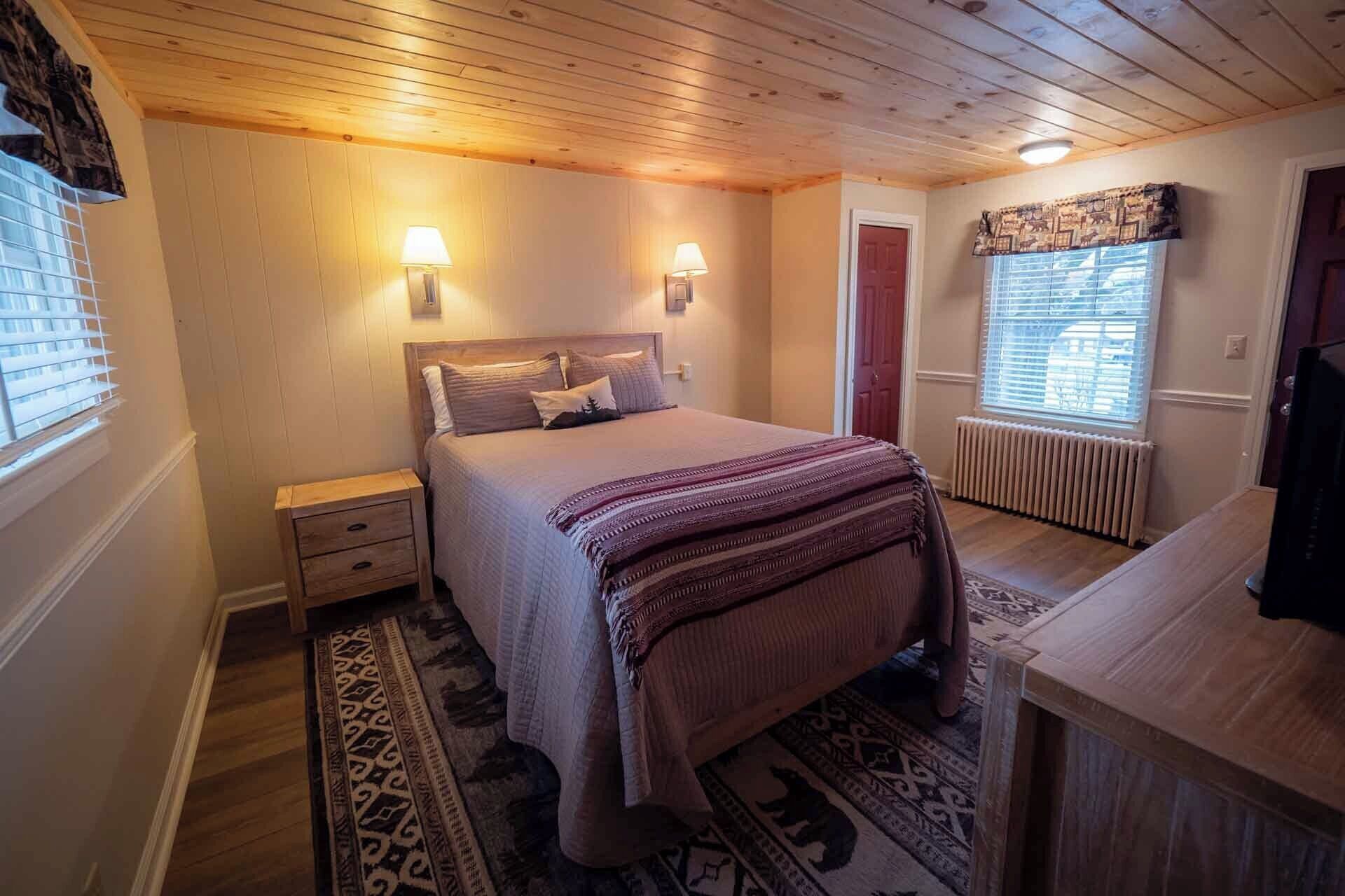 A bedroom with a wooden ceiling, a bed with a patterned blanket, bedside lamps, a radiator, and a window with blinds.