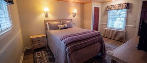 A bedroom with a wooden ceiling, a bed with a patterned blanket, bedside lamps, a radiator, and a window with blinds.