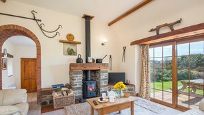 A cozy living room with a stone fireplace, a television, and a view of the garden through large windows. - Kingfisher Cottage, Holiday Cottage St Ishmaels, Near Dale, Sleeps 4, 2 bedrooms,  bathrooms (Haverfordwest)
