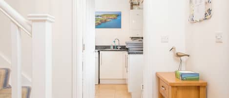 A hallway leading to a kitchen with a view of the sea, a wooden table with a lamp, and a wall-mounted calendar.