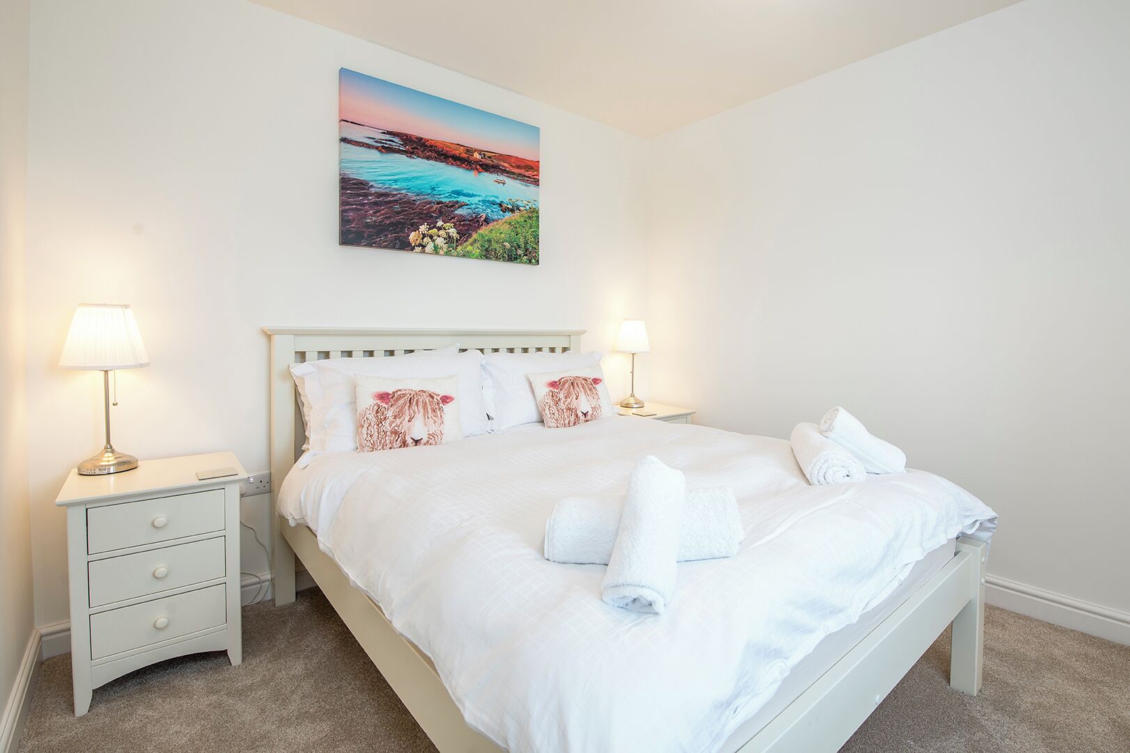 A neatly made bed with white linens and two pillows, a bedside table with a lamp, and a painting of a coastal landscape above the bed.