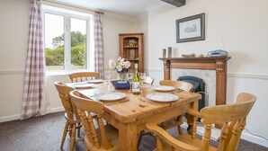 A dining room with a wooden table set for six, surrounded by wooden chairs. A fireplace with a mantel is visible in the background, and a small wooden cabinet is placed against the wall. - Little Mead, Holiday Cottage Amroth, Sleeps 4, 2 bedrooms,  bathrooms (Amroth)