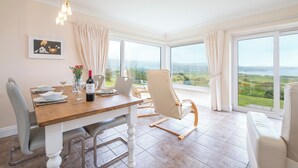 Un coin repas avec une table en bois, des chaises blanches et une vue sur la campagne, grâce à de grandes fenêtres.