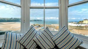 A room with a large window overlooking a beach and sea, featuring striped pillows and a view of boats and buildings.
